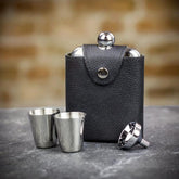A black leather hip flask set with two stainless steel cups and a funnel, displayed on a stone surface.