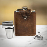 A brown leather hip flask with a stainless steel cap and two matching cups, displayed on a marble surface.