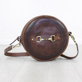 A round brown leather handbag with a metallic bit decoration in the center, fitted with a shoulder strap and a zip closure.