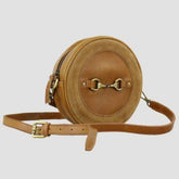 A tan leather and suede bag with a round design, featuring a zip closure, snaffle fitment, and an adjustable leather strap.