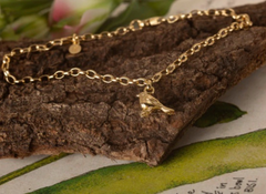 Gold chain bracelet with a bird charm on a textured surface with green leaves.