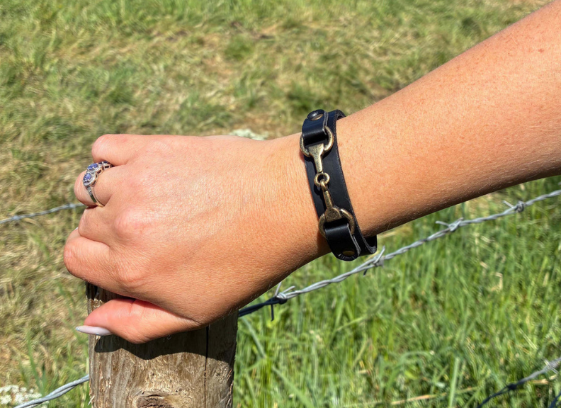 Hand wearing a black leather bracelet holding onto a barbed wire fence with grass in the background