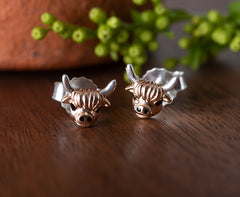 Pair of bull-shaped earrings on a wooden surface with greenery in the background