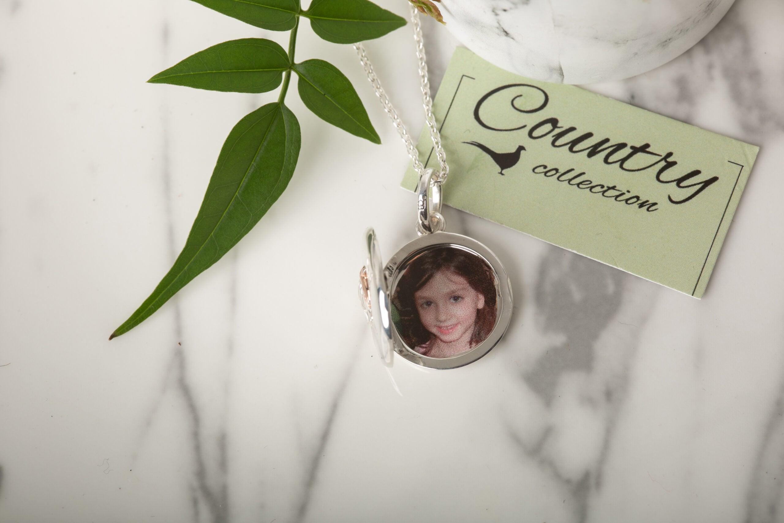 Silver locket with a photo of a young girl on a marble surface with a 'Country Collection' tag.