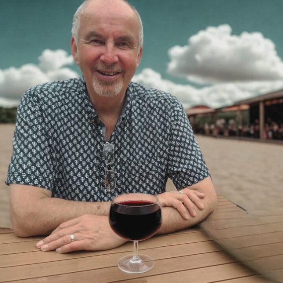 Man sitting at a wooden table with a glass of red wine, outdoors with clouds in the background